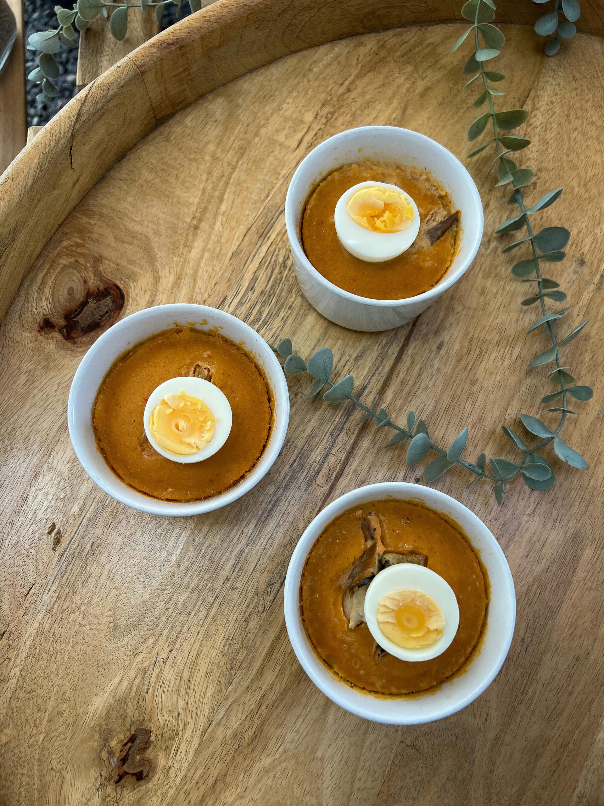 Three ramekins with savory egg dishes on a wooden tray, garnished with eucalyptus.